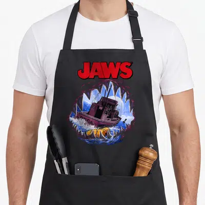 Mouth movie poster apron - JAWS text above cartoon shark jaws framing a purple boat and yellow buoy on blue water — shown on a black cotton canvas apron against a white background