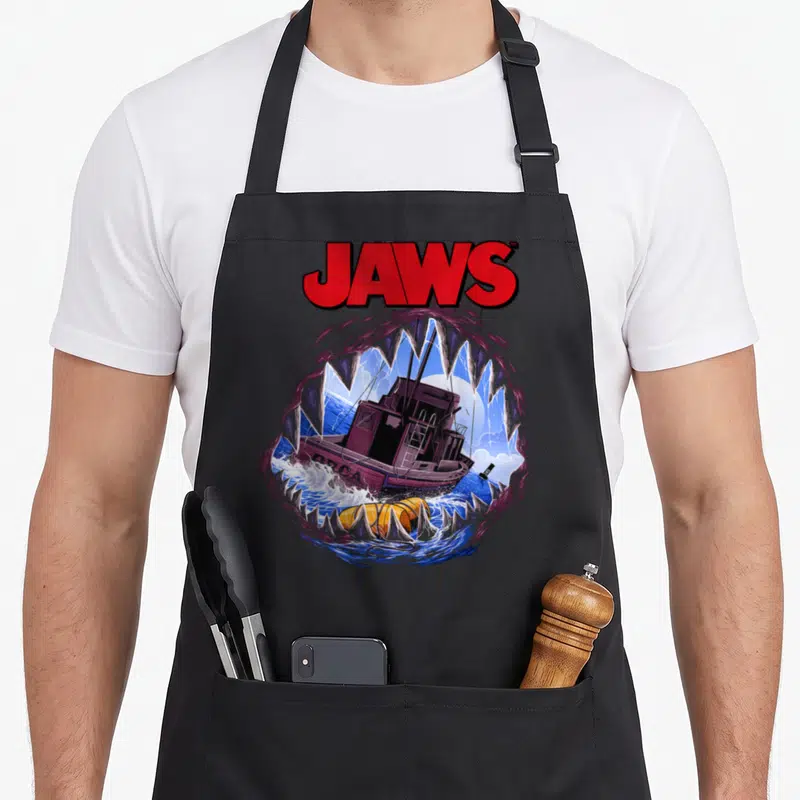 Mouth movie poster apron - JAWS text above cartoon shark jaws framing a purple boat and yellow buoy on blue water — shown on a black cotton canvas apron against a white background