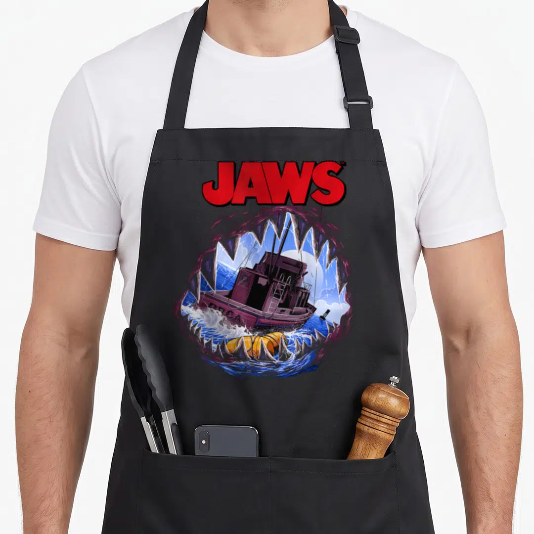 Mouth movie poster apron - JAWS text above cartoon shark jaws framing a purple boat and yellow buoy on blue water — shown on a black cotton canvas apron against a white background