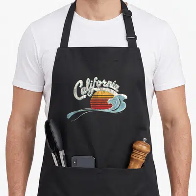 Surfer 70s apron - Arched California text over striped sunset and blue wave in retro style — shown on a black cotton canvas apron against a white background