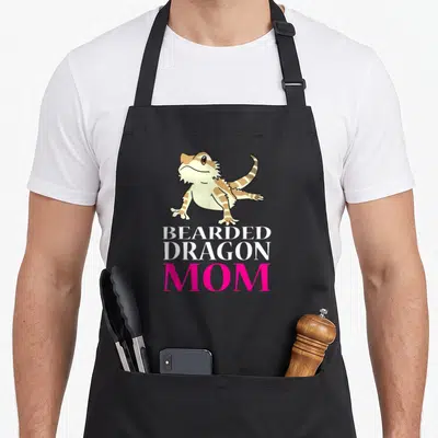 Beared pet apron - Cartoon bearded dragon above stacked text reading BEARDED DRAGON MOM in white and pink — shown on a black cotton canvas apron against a white background