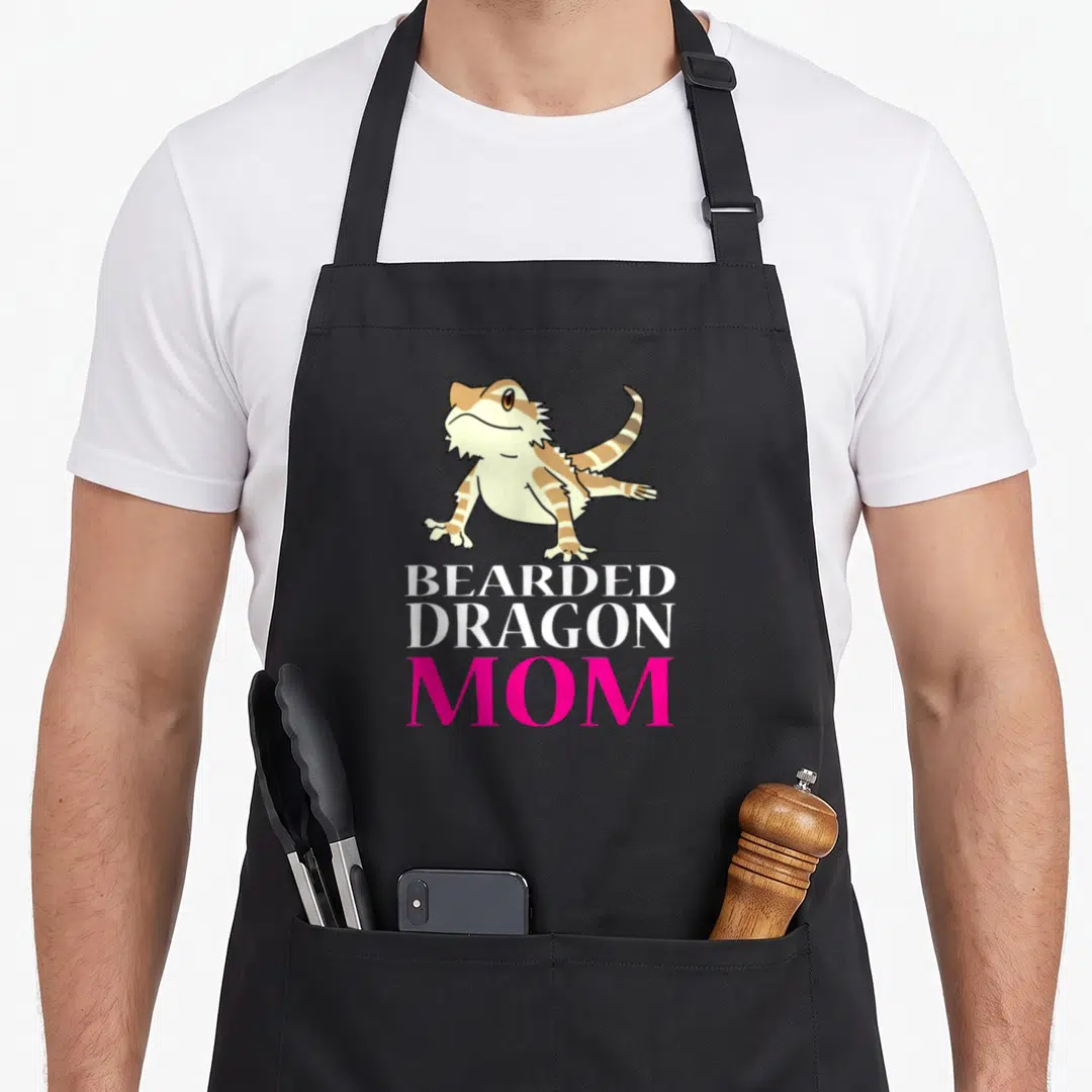 Beared pet apron - Cartoon bearded dragon above stacked text reading BEARDED DRAGON MOM in white and pink — shown on a black cotton canvas apron against a white background
