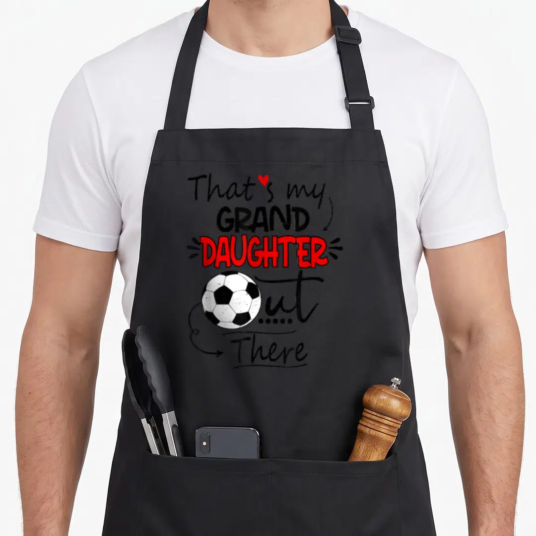Granddaughter grandma aprons - Stacked text That's my Grand Daughter Out There with soccer ball and red heart — shown on a black cotton canvas apron against a white background