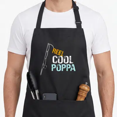 Poppa fishing apron - Fishing rod graphic next to stacked Reel Cool Poppa text in orange, white, and blue distressed — shown on a black cotton canvas apron against a white background