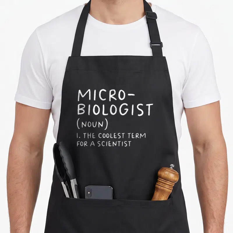 Microbiologist teacher apron - White stacked text defining microbiologist as the coolest term for a scientist — shown on a black cotton canvas apron against a white background