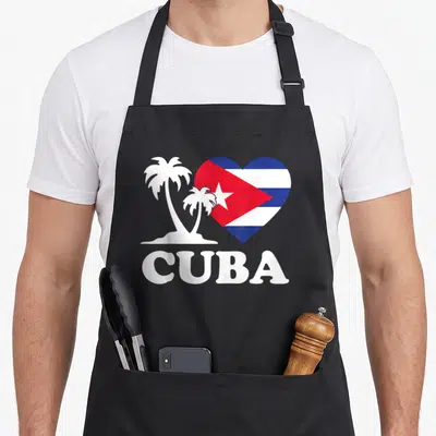 Cuba flag cuban apron - Heart with Cuban flag stripes and star next to two palm trees on sand with CUBA text below — shown on a black cotton canvas apron against a white background