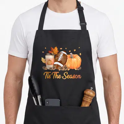 Football fall apron - Tis The Season script below watercolor football, coffee cup, pumpkin, leaves, and pinecones — shown on a black cotton canvas apron against a white background