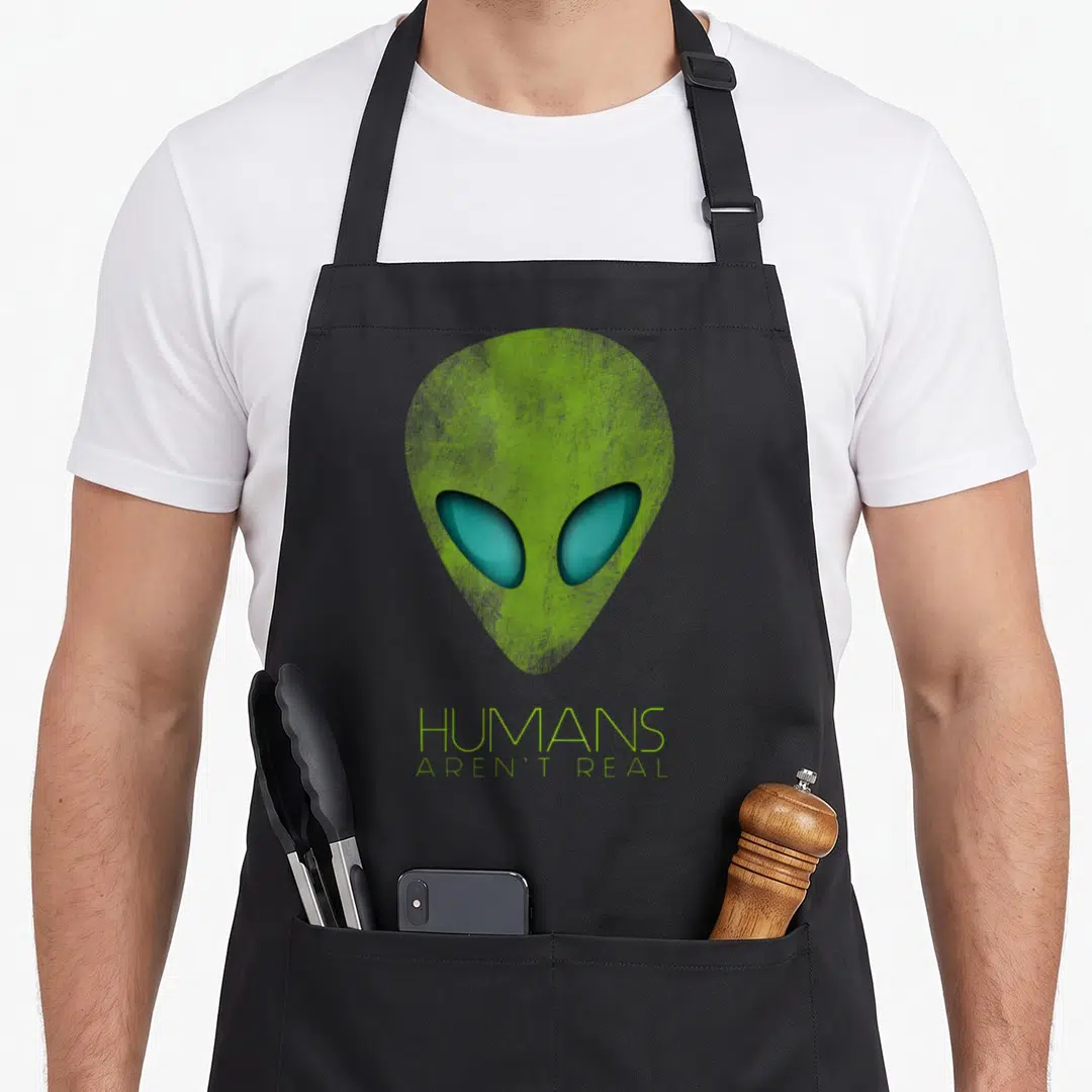 Human alien ufo apron - Lime green alien head with teal eyes above HUMANS AREN'T REAL text — shown on a black cotton canvas apron against a white background