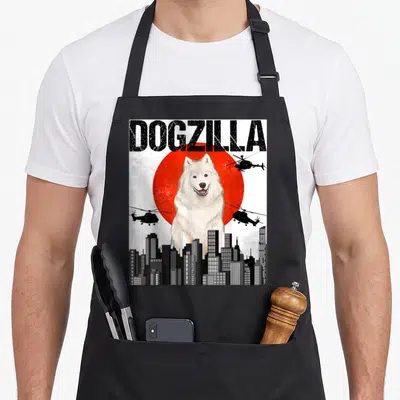 Dogzilla samoyed apron - Giant white dog in front of red circle with helicopters and skyline under DOGZILLA text — shown on a black cotton canvas apron against a white background