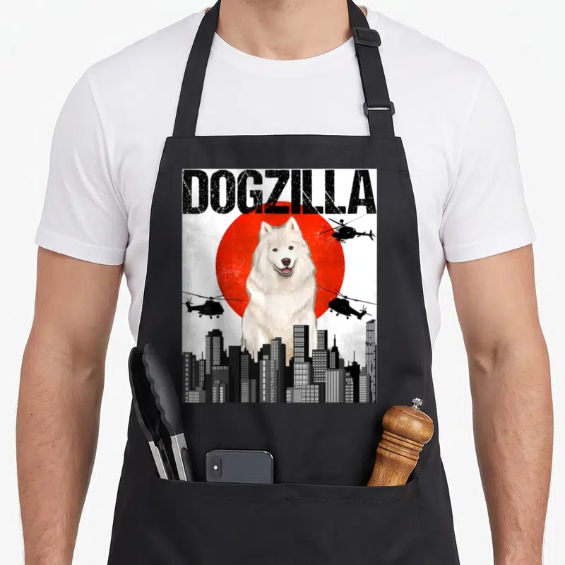 Dogzilla samoyed apron - Giant white dog in front of red circle with helicopters and skyline under DOGZILLA text — shown on a black cotton canvas apron against a white background
