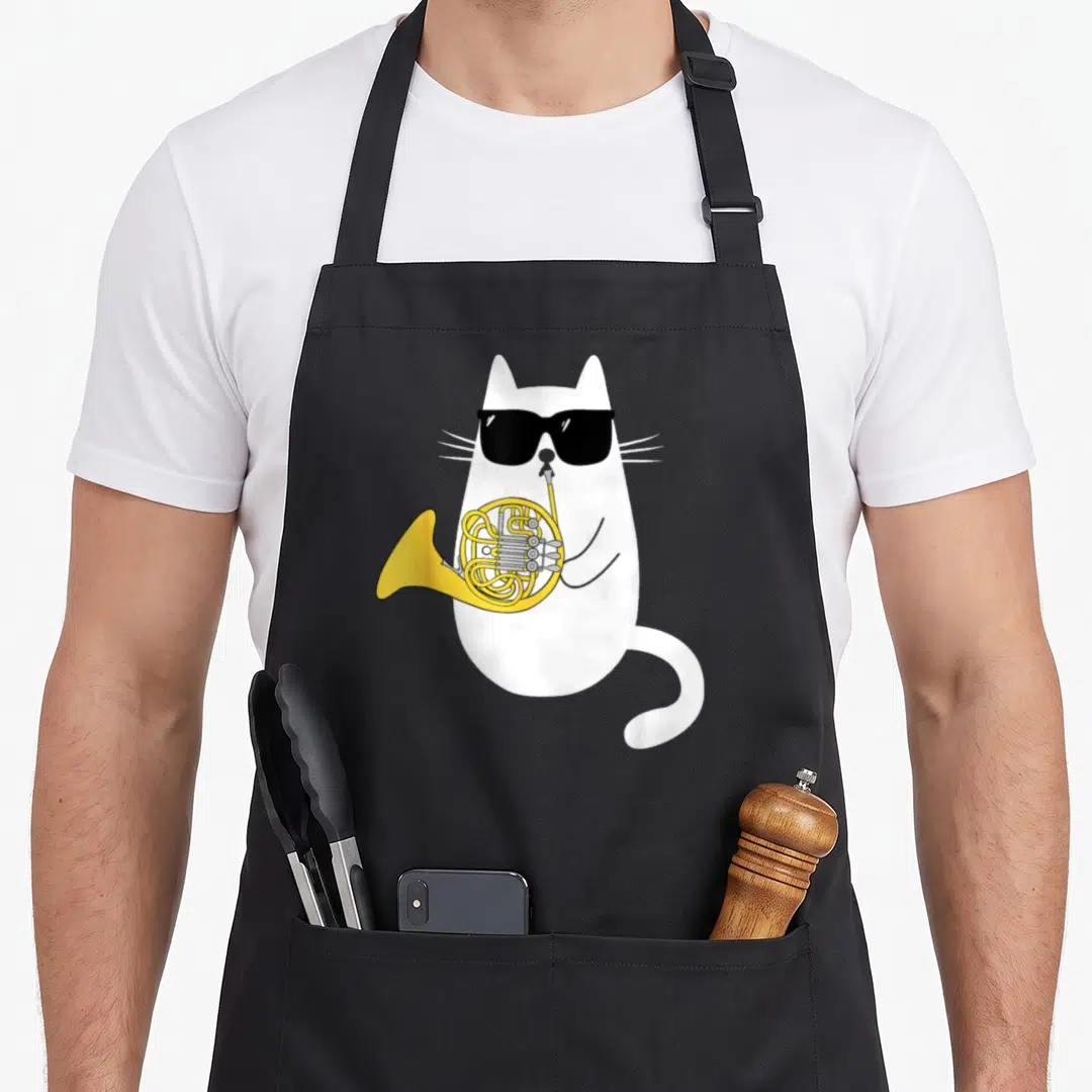 Horn cute cat apron - White cartoon cat wearing black sunglasses playing a yellow french horn — shown on a black cotton canvas apron against a white background