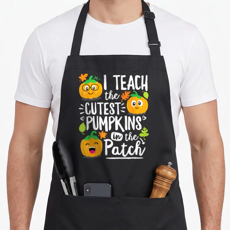 Cutest teacher apron - Three smiling orange cartoon pumpkins with white text I TEACH the CUTEST PUMPKINS in the Patch — shown on a black cotton canvas apron against a white background