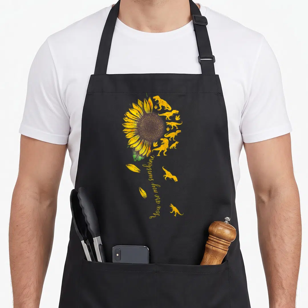 Sunshine sunflower apron - Yellow dinosaurs forming a sunflower stem with You are my sunshine text and floating petals — shown on a black cotton canvas apron against a white background