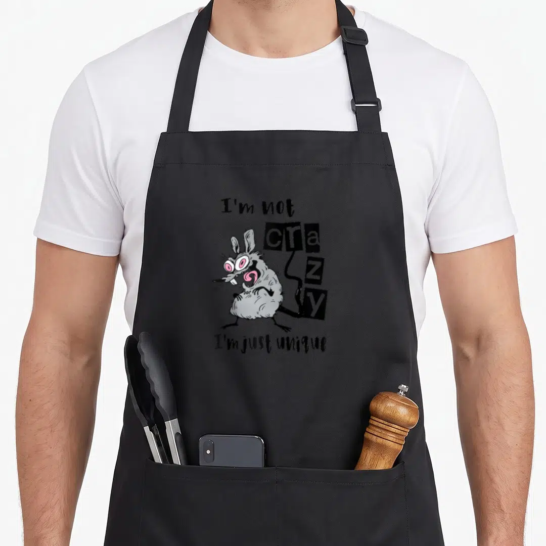 Weird unique apron - Cartoon grey mouse with pink eyes next to text I'm not crazy I'm just unique — shown on a black cotton canvas apron against a white background