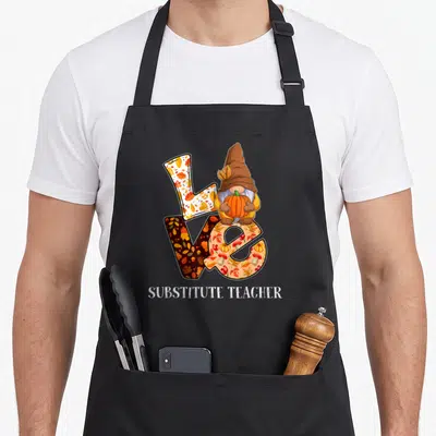 Autumn teacher apron - Gnome holding pumpkin sits on patterned LOVE letters with SUBSTITUTE TEACHER text below — shown on a black cotton canvas apron against a white background