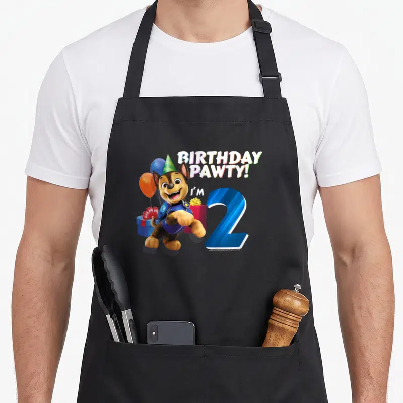Retro chase birthday apron - Stacked text BIRTHDAY PAWTY I'M 2 with a brown rescue pup in a blue vest and party hat — shown on a black cotton canvas apron against a white background