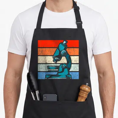 Microscope laboratory apron - Blue microscope centered on five horizontal stripes in red, orange, beige, light blue — shown on a black cotton canvas apron against a white background