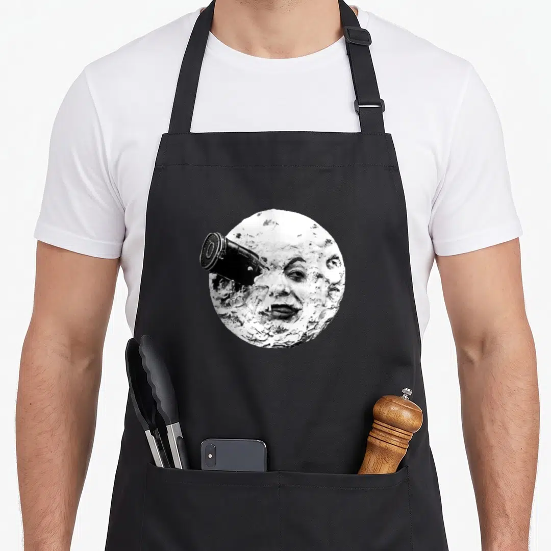 Trip moon vintage apron - Black and white moon with human face and protruding camera lens on left side — shown on a black cotton canvas apron against a white background