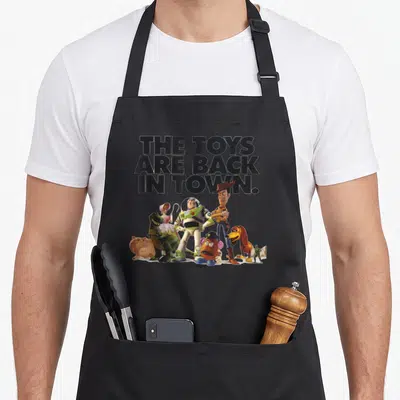 Toy camping apron - Stacked text THE TOYS ARE BACK IN TOWN above a cluster of cartoon figures in black white green blue — shown on a black cotton canvas apron against a white background