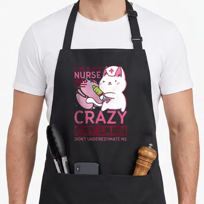 Nurse crazy cat lady apron - Cartoon white cat holding green syringe with pink and maroon text on black background — shown on a black cotton canvas apron against a white background