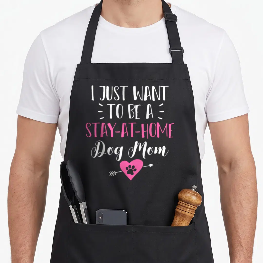 Puppy at home aprons - I Just Want to be a Stay-at-Home Dog Mom text above pink heart with white arrow and paw print — shown on a black cotton canvas apron against a white background