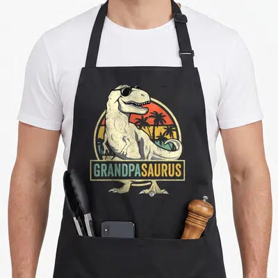 Grandpasaurus t-rex apron - T-Rex in sunglasses before retro sunset with GRANDPASAURUS banner text — shown on a black cotton canvas apron against a white background