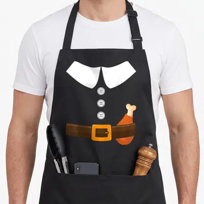 Leg pilgrim apron - Cartoon white collar, three buttons, brown belt with orange buckle, and turkey leg tucked — shown on a black cotton canvas apron against a white background