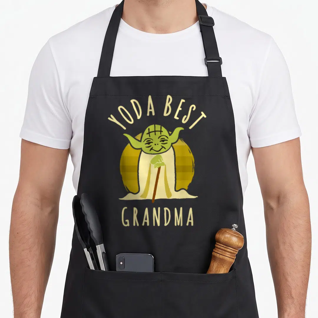 Yoda grandma apron - Cartoon Yoda with cane inside striped yellow circle, beige YODA BEST GRANDMA text — shown on a black cotton canvas apron against a white background
