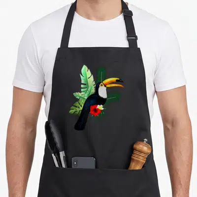 Toucan the floral apron - Realistic toucan with yellow beak perched on green tropical leaves above a red flower — shown on a black cotton canvas apron against a white background
