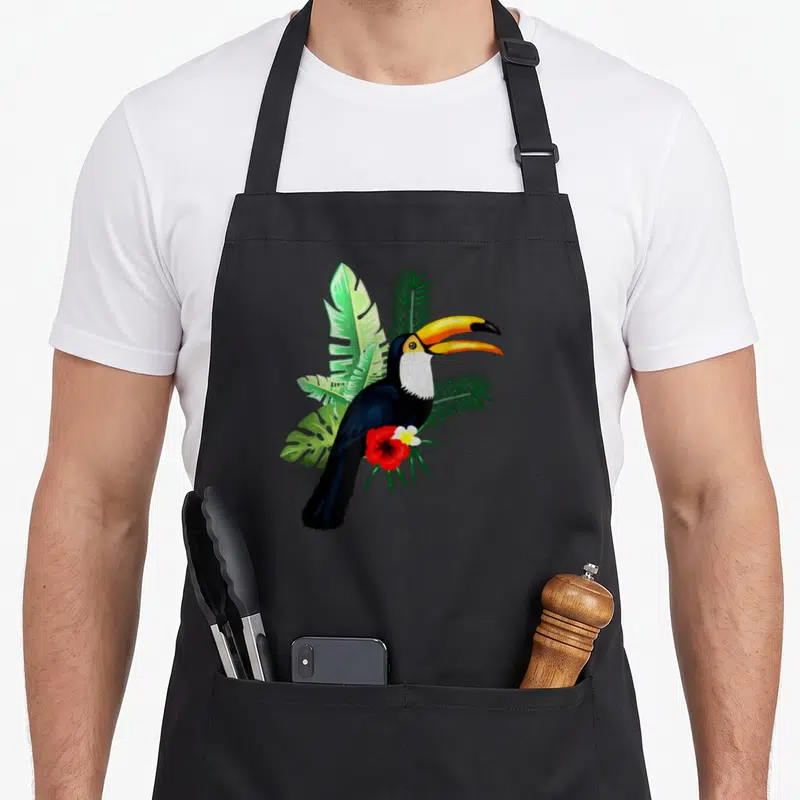 Toucan the floral apron - Realistic toucan with yellow beak perched on green tropical leaves above a red flower — shown on a black cotton canvas apron against a white background