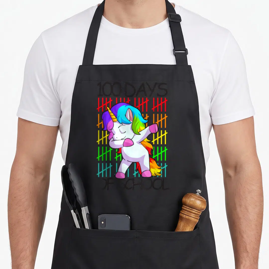 School unicorn apron - Cartoon unicorn dabbing in front of rainbow tally marks with 100 DAYS OF SCHOOL text — shown on a black cotton canvas apron against a white background