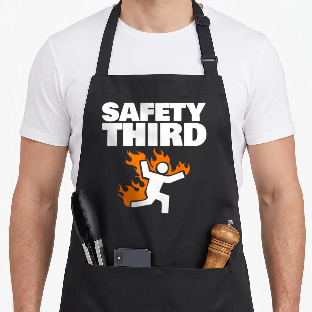 Cartoon safety apron - SAFETY THIRD text above running stick figure on fire in orange and white — shown on a black cotton canvas apron against a white background