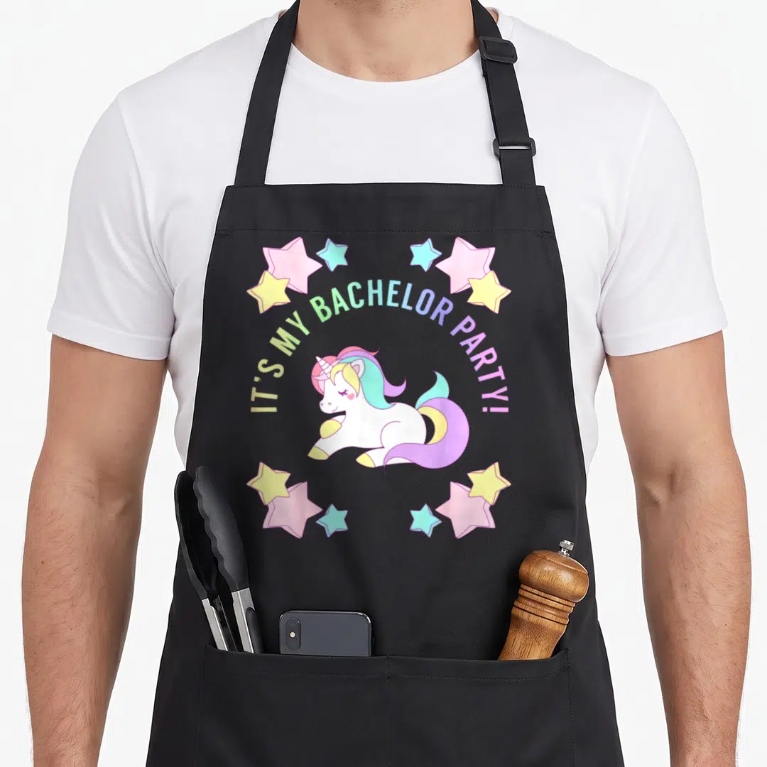 Funny bachelor apron - Sitting unicorn with rainbow mane under arched text surrounded by pastel stars — shown on a black cotton canvas apron against a white background