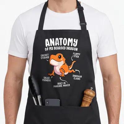 Bearded anatomy apron - Cartoon orange lizard with white arrows labeling body parts as cricket storage and dinosaur — shown on a black cotton canvas apron against a white background