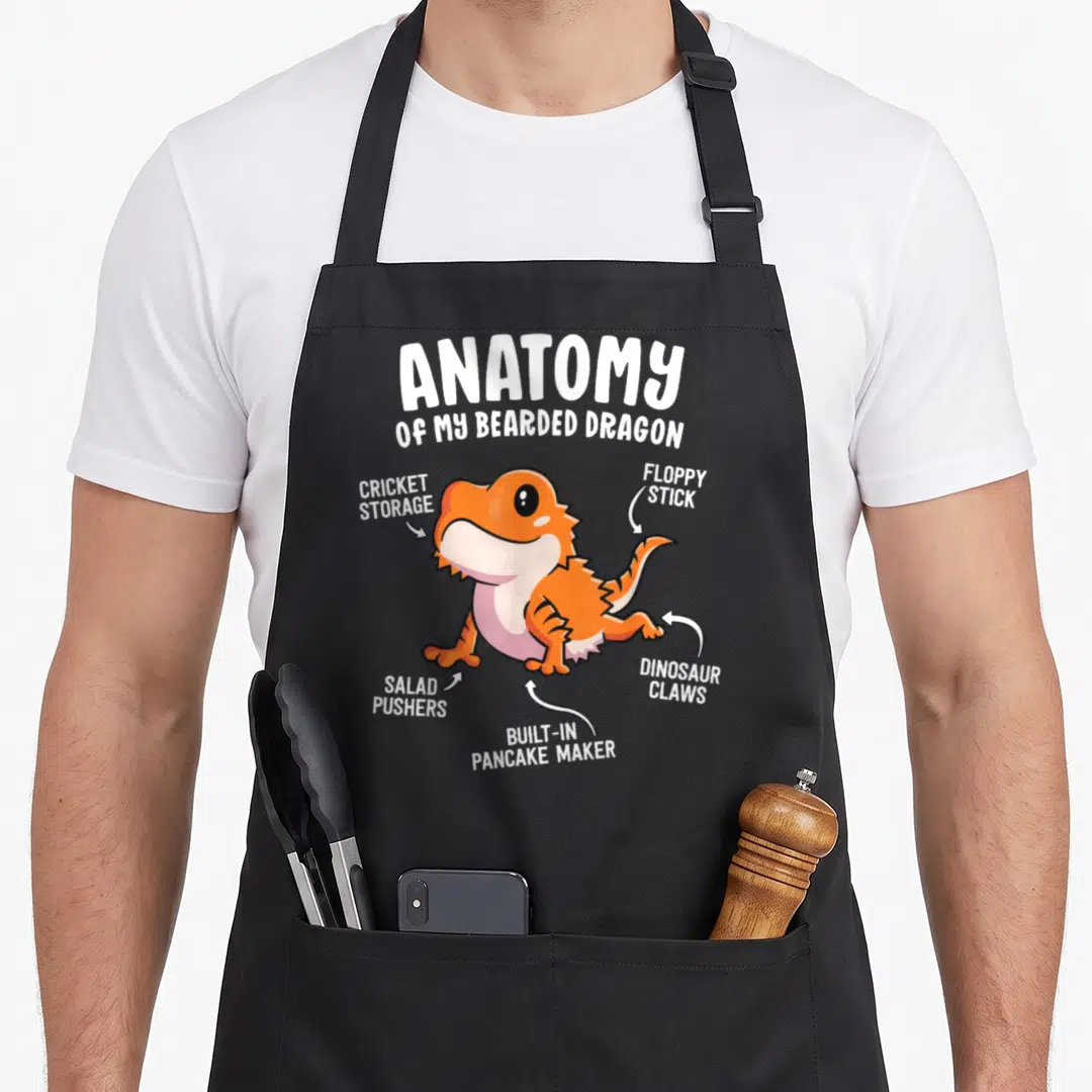 Bearded anatomy apron - Cartoon orange lizard with white arrows labeling body parts as cricket storage and dinosaur — shown on a black cotton canvas apron against a white background