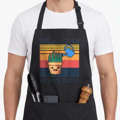 Face plant daddy apron - Cactus in a pot being watered by a blue can with text YES ALL OVER MY FACE DADDY on striped — shown on a black cotton canvas apron against a white background