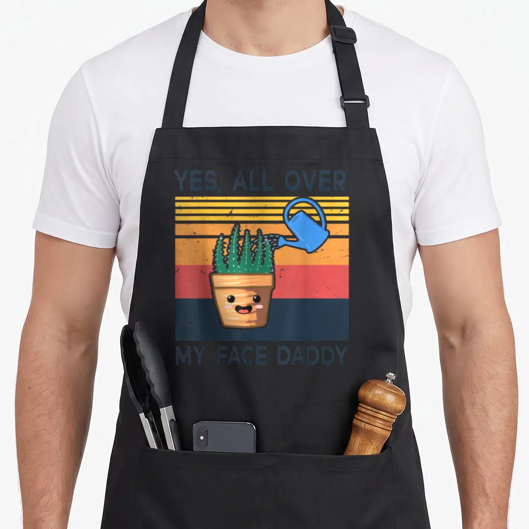 Face plant daddy apron - Cactus in a pot being watered by a blue can with text YES ALL OVER MY FACE DADDY on striped — shown on a black cotton canvas apron against a white background