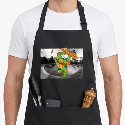 Skating michelangelo apron - Green cartoon turtle handstanding with nunchucks and skateboard over grey skate bowl — shown on a black cotton canvas apron against a white background