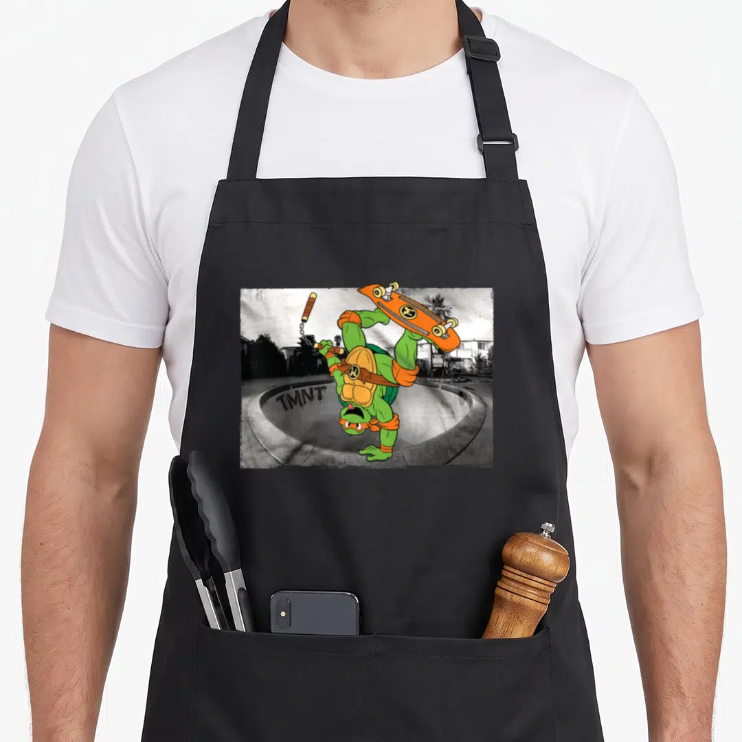 Skating michelangelo apron - Green cartoon turtle handstanding with nunchucks and skateboard over grey skate bowl — shown on a black cotton canvas apron against a white background