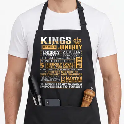 Birthday january king apron - Stacked white and orange text listing ten traits for January kings with crown icon — shown on a black cotton canvas apron against a white background