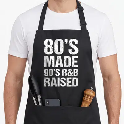 School r aprons - White distressed stacked text reading 80s made 90s R&B raised — shown on a black cotton canvas apron against a white background