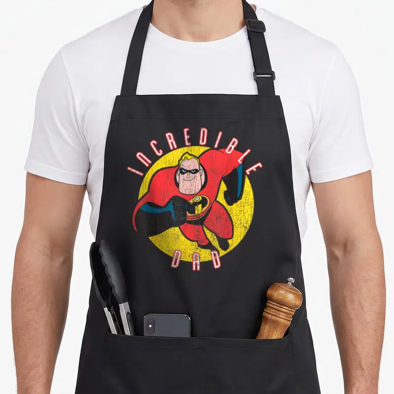 Funny incredible father day apron - Distressed INCREDIBLE DAD text around yellow circle with red suited superhero figure — shown on a black cotton canvas apron against a white background