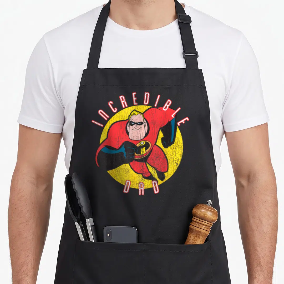 Funny incredible father day apron - Distressed INCREDIBLE DAD text around yellow circle with red suited superhero figure — shown on a black cotton canvas apron against a white background