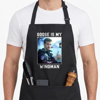 Wingman goose apron - Man in flight suit thumbs up with GOOSE IS MY WINGMAN text — shown on a black cotton canvas apron against a white background
