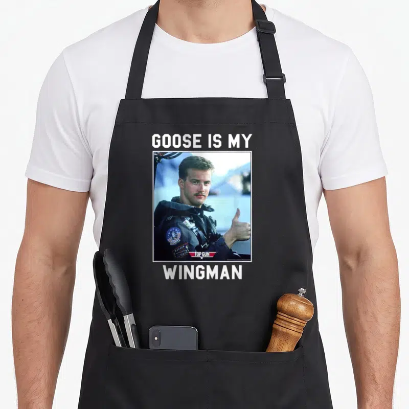 Wingman goose apron - Man in flight suit thumbs up with GOOSE IS MY WINGMAN text — shown on a black cotton canvas apron against a white background