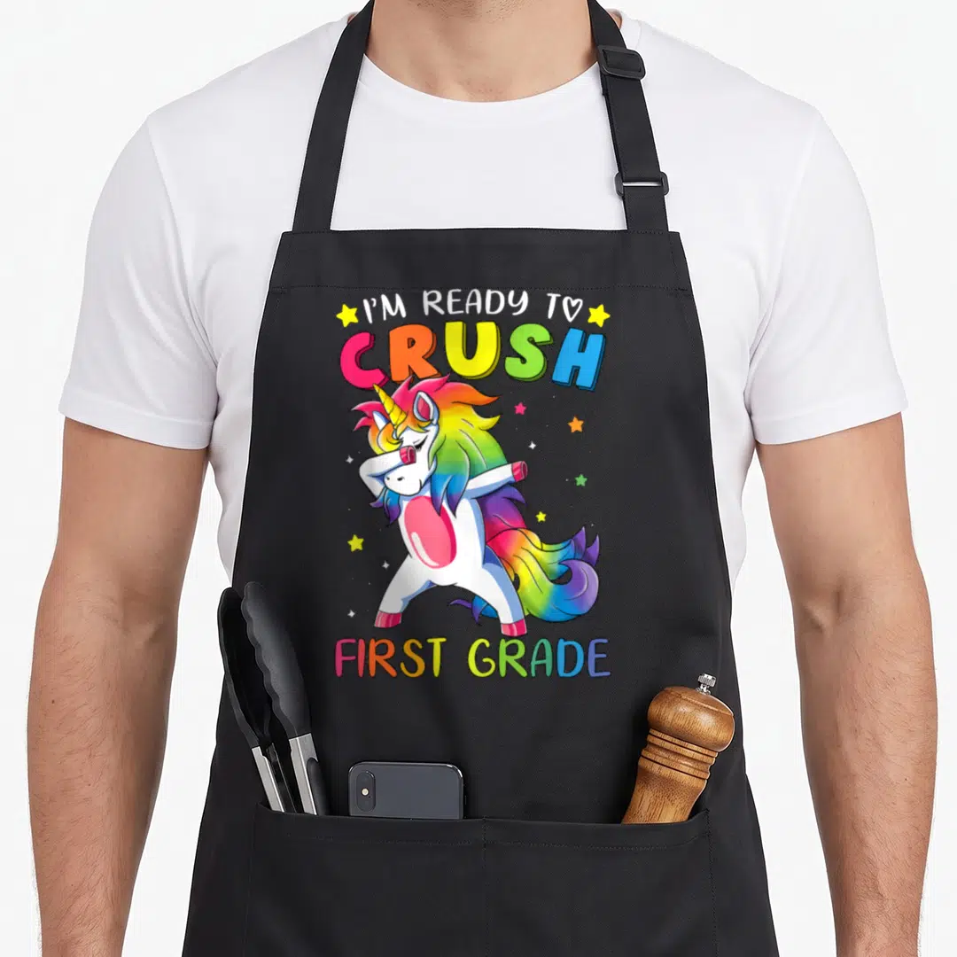 Ready unicorn apron - Dabbing rainbow unicorn with stars and text I'M READY TO CRUSH FIRST GRADE — shown on a black cotton canvas apron against a white background