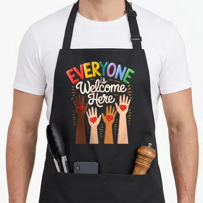 Welcome idaho teacher apron - Four diverse hands with red hearts reaching up under rainbow text reading EVERYONE is — shown on a black cotton canvas apron against a white background