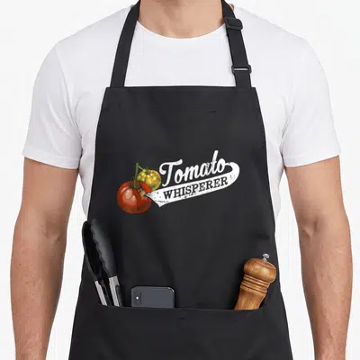 Gardening tomato apron - Two red and yellow tomatoes with stem beside Tomato WHISPERER text in distressed white banner — shown on a black cotton canvas apron against a white background