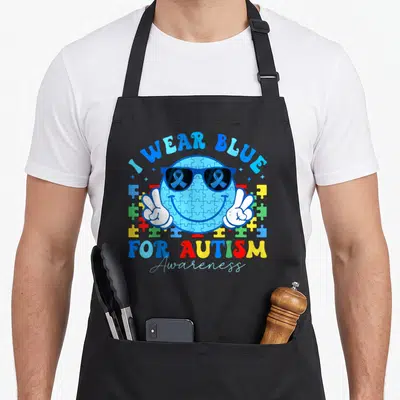 Month teacher art apron - Blue puzzle smiley face with sunglasses, white peace hands, and I WEAR BLUE text surrounded — shown on a black cotton canvas apron against a white background