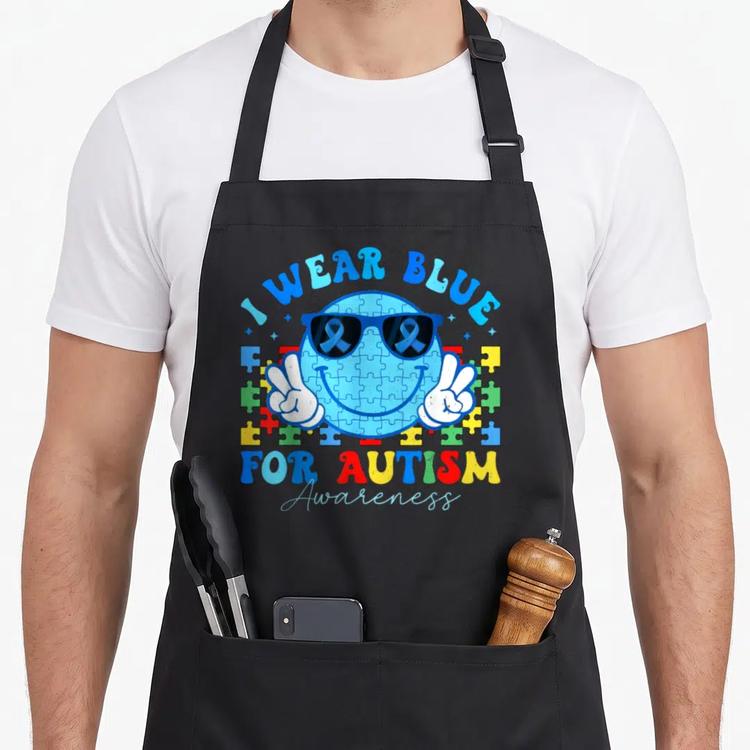 Month teacher art apron - Blue puzzle smiley face with sunglasses, white peace hands, and I WEAR BLUE text surrounded — shown on a black cotton canvas apron against a white background